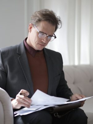 Elegant adult man in jacket and glasses looking through documents while sitting on white sofa in luxury room