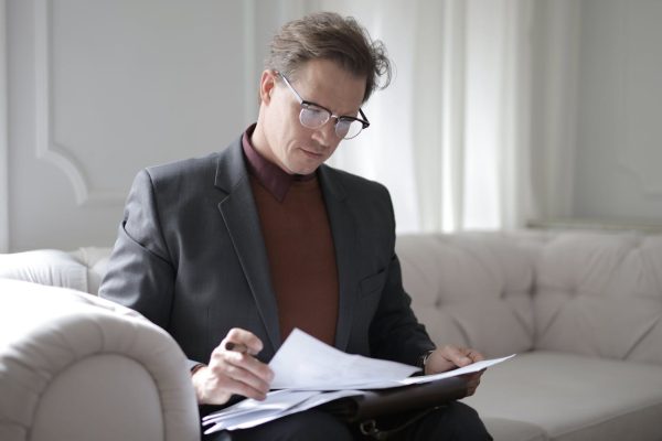 Elegant adult man in jacket and glasses looking through documents while sitting on white sofa in luxury room